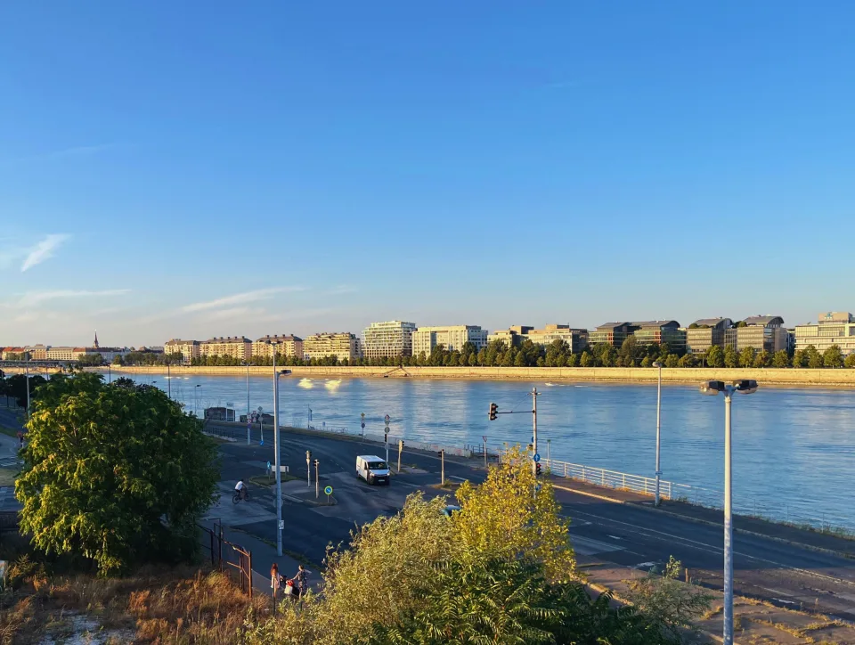 Duna-parti városrész Budapesten, modern irodaházakkal és lakóépületekkel, kék ég alatt, a rakparti úton autók és biciklisek láthatók.